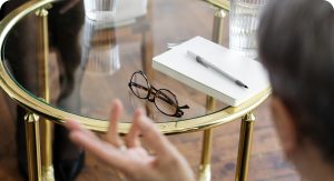 Скляний круглий стіл з золотистою рамою, на якому лежать окуляри в коричневій оправі, блокнот з ручкою та склянка води, у передньому плані - розмиті руки людини і частина голови.