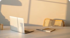 Робочий стіл з відкритим ноутбуком, блокнотом і склянкою, на задньому фоні стілець з плетеною спинкою, освітлений м'яким теплим світлом ранкового або вечірнього сонця.