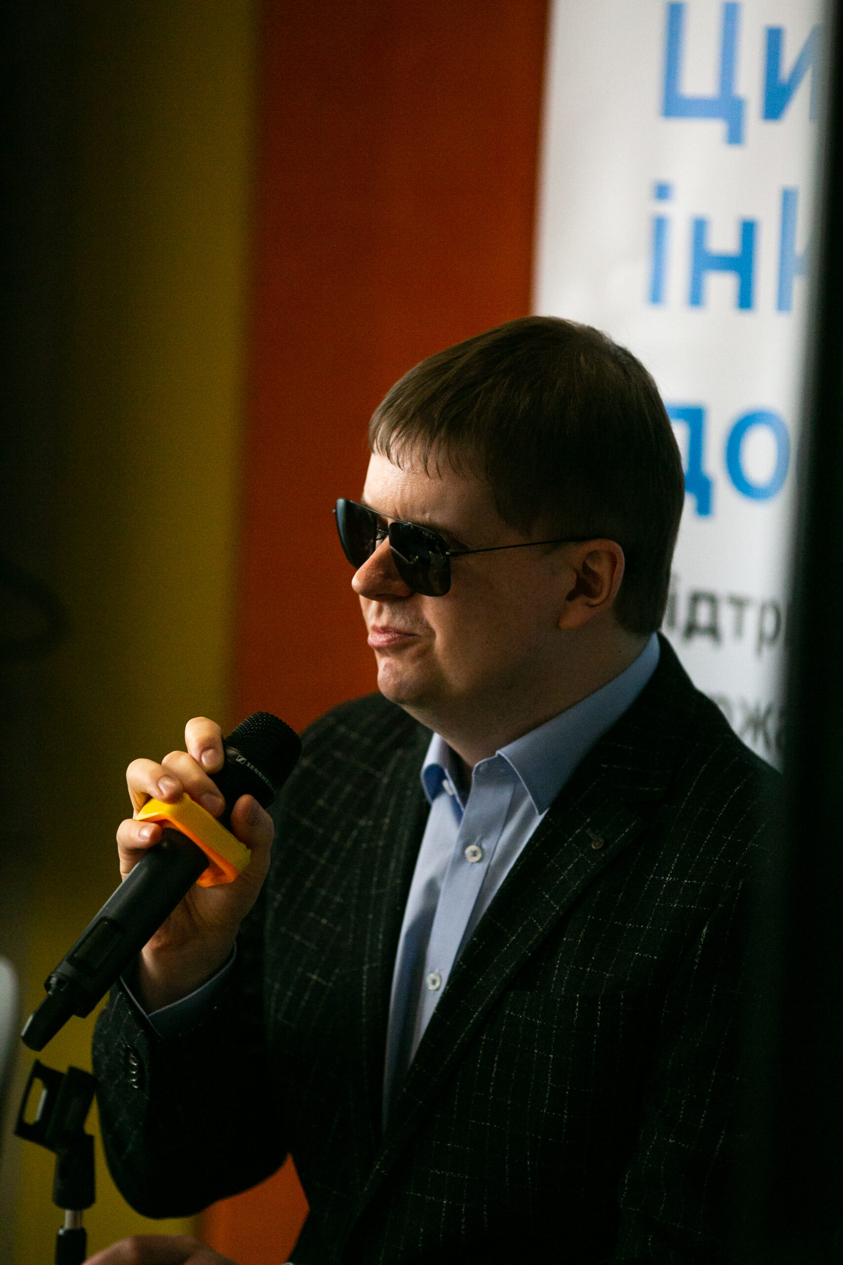 Чоловік у темних окулярах, блакитній сорочці та темному піджаку тримає мікрофон у руці на фоні розмитого плаката з текстом синім кольором.