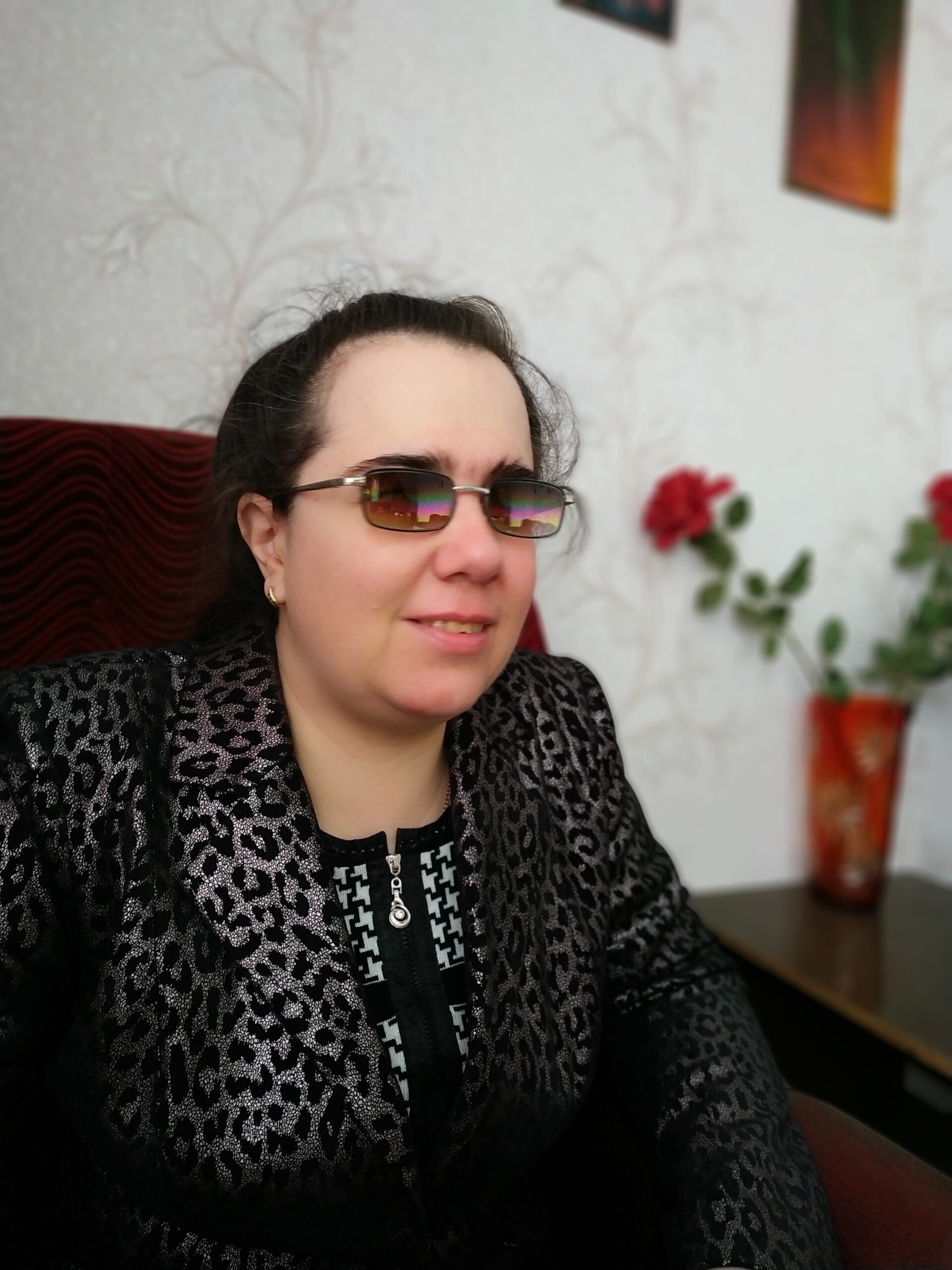 Жінка в темних окулярах із темним зібраним волоссям, одягнена в блискучий жакет із леопардовим візерунком. На тлі — світла стіна, вазон із червоною квіткою та картини