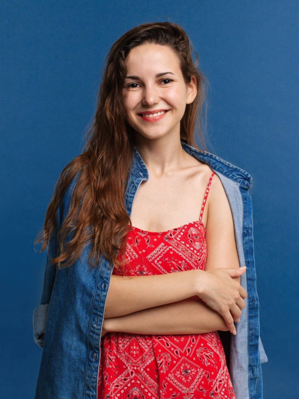 Усміхнена молода жінка з довгим хвилястим каштановим волоссям, одягнена у червону сукню з білим орнаментом і джинсову сорочку