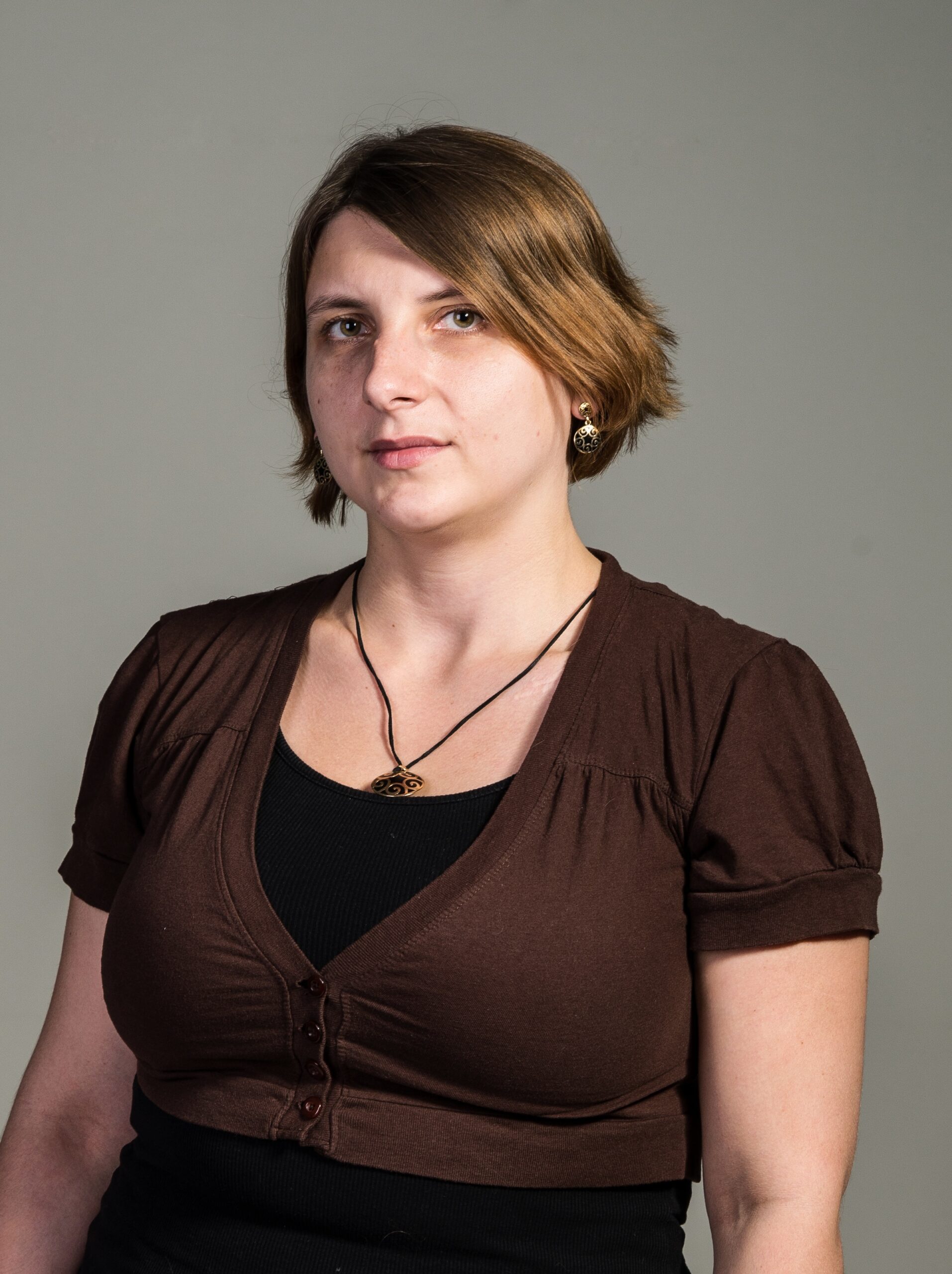 Жінка зі світло-коричневим коротким волоссям, одягнена в чорний топ і коричневу кофту, на одязі та сережках прикраси із дерева з візерунками, на сірому тлі.